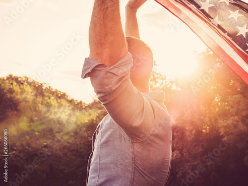 Handsome man waving an American flag against a background of trees and blue sky. View from the back, close-up. National holiday concept