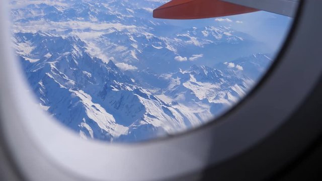 Aerial view of European Alps while flying over snow covered mountains. Showing porthole frame and aircraft wing camera slowly moving in to get clear view of  mountain peaks