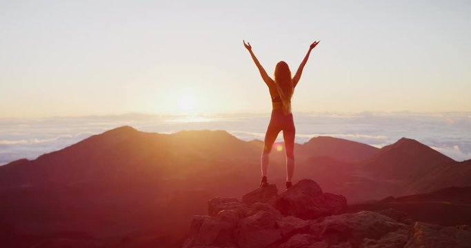 Young athletic woman raising her arms into the air in success pose while she looks out at epic sunrise from a mountaintop, amazing outdoors fitness in slow motion
