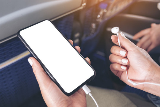 Mockup image of woman's hand holding a black smart phone with blank desktop screen and earphone in cabin