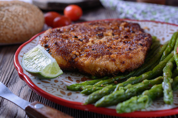 Hot appetizing schnitzel with crispy crust on a plate next to asparagus, lime and cherry tomatoes on a wooden table are ready for eating