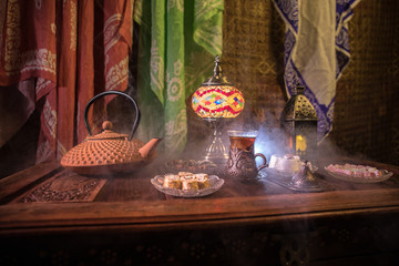 Arabian tea in glass with eastern snacks on vintage wooden surface. Eastern tea concept. Low light lounge interior with carpet. Empty space.