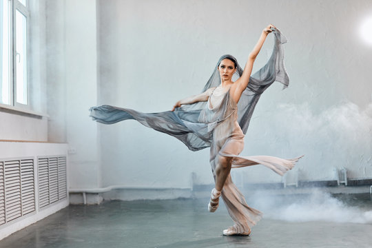 Female ballet dancer with white flowing fabric. Flow shapes and movement in the studio on white smoky background.
