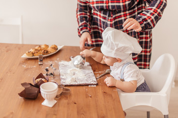 Fototapeta na wymiar Adorable little baker covered with flour, wearing white Chef s hat, playing with Mom s kitchen tableware sitting with spoon in hands at table.