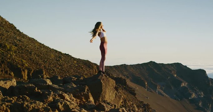 Young athletic woman standing on the edge of a cliff with her arms in the air watching an epic sunrise, amazing outdoors fitness lifestyle