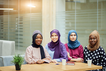 Group of beautiful young muslim business women wearing hijabs of different colour, are sharing info, sitting together at table in business centre