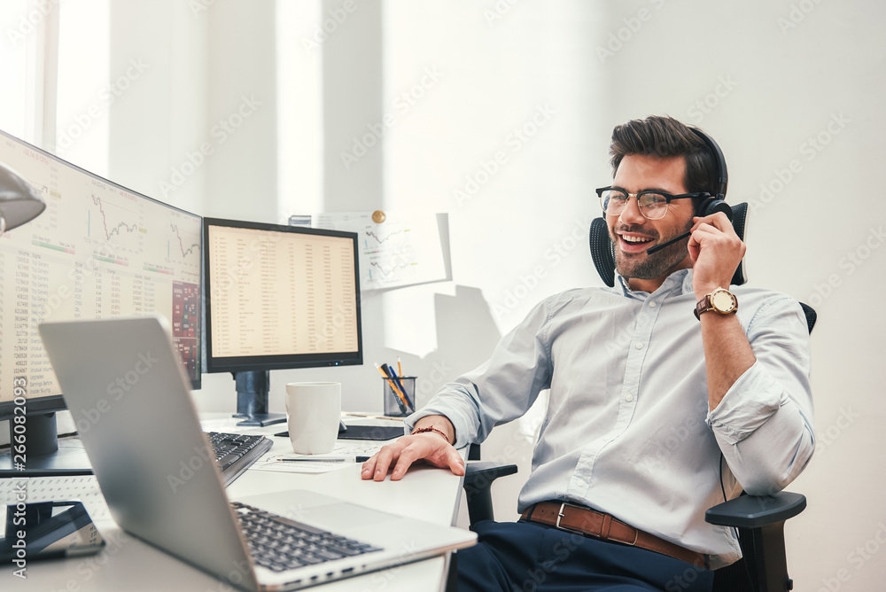 Poster good news. happy young bearded trader in headset talking with client and smiling while sitting in hi
