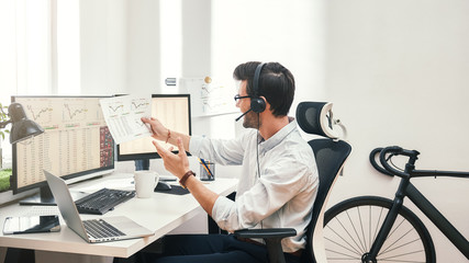 Growing chart. Side view of happy young bearded trader in headset holding financial report, talking with client and smiling while sitting in front of monitor screens in the office.