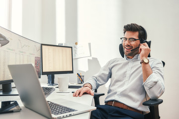 Good news. Happy young bearded trader in headset talking with client and smiling while sitting in...