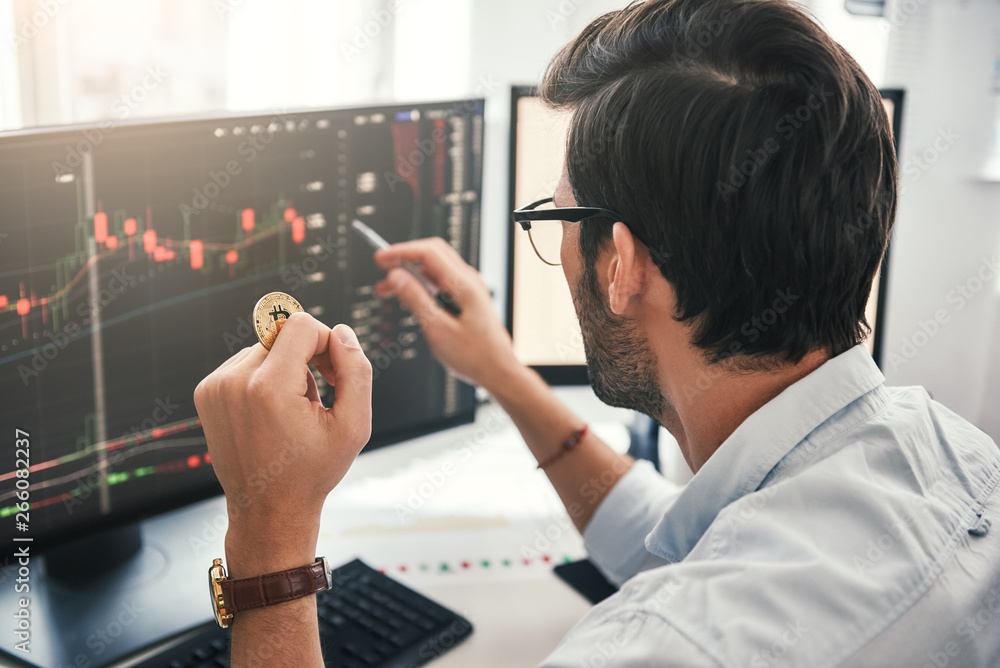 Fototapete Exchange stock market. Back view of young professional trader in eyeglasses pointing on the data on computer screen with pen and holding Bitcoin in one hand while working his modern office.