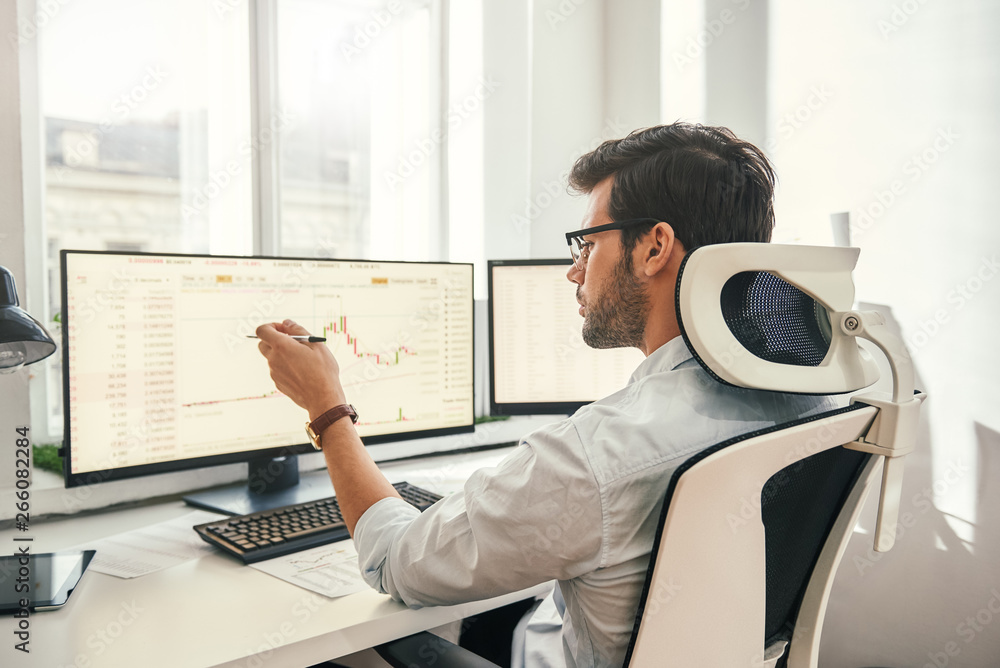 Fototapete analyzing data. back view of young businessman or trader pointing on the data on computer screen wit