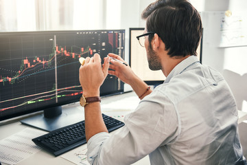 Successful trading. Back view of young bearded trader in formal wear pointing on the data on...
