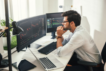 Successful trader. Back view of bearded stock market broker in eyeglasses analyzing data and graphs on multiple computer screens while sitting in modern office. - Powered by Adobe