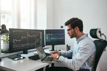 Workplace of trader. Young bearded trader wearing eyeglasses using his laptop while sitting in office in front of computer screens with trading charts and financial data