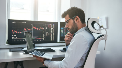 Busy day. Professional male trader wearing eyeglasses analyzing financial market via laptop while sitting in front of computer screens with trading charts in modern office interior