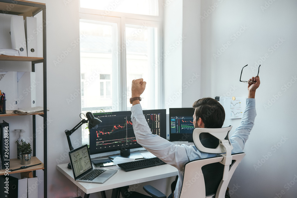 Poster winner! back view of stock trader with raised hands holding eyeglasses and feeling happy while looki