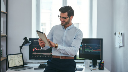 Good news. Happy young trader in formal wear is looking is using his digital tablet and smiling while standing in front of computer screens with trading charts in the office. - obrazy, fototapety, plakaty