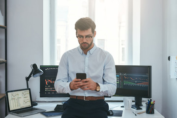Modern technologies. Successful young trader in formal wear and eyeglasses using smartphone while standing at office in front of computer screens with trading charts