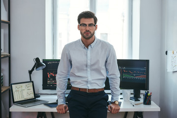 Modern trader. Successful young businessman in formal clothes and eyeglasses looking at camera while standing in the office