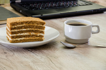 a piece of cake on a plate and a cup of coffee on the background of a laptop and notebook
