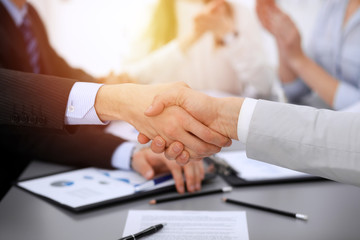 Business people shaking hands at meeting while theirs colleagues clapping and applauding. Group of unknown businessmen and women in modern white office. Success teamwork, partnership and handshake