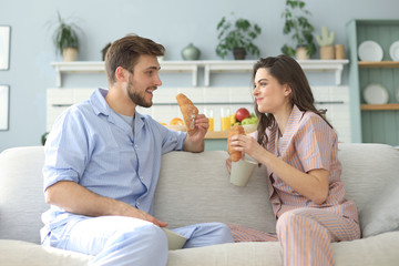 Happy young couple in pajamas in kitchen having breakfast, feeding each other a croissant.