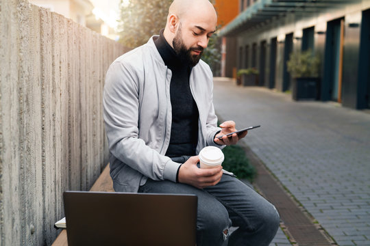 Man sits on bench and uses smartphone during coffee break,next is laptop.Guy checks email on mobile phone, sends text message.