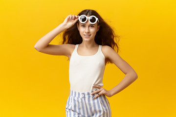 Pretty girl in her twenties posing at yellow studio wall wearing white top, striped trousers and stylish sunglasses in round frame, taking off eyewear to see clearly. People, lifestlye and fashion