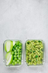 Vegetarian meal prep containers green pesto pasta with basil and pine nuts. Apple and grape for dessert. Top view