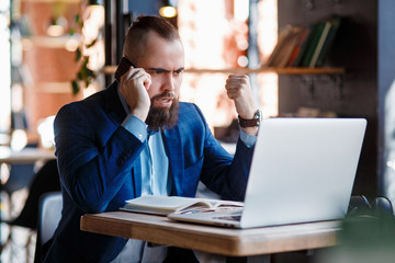 Obraz premium Serious bearded businessman conducts telephone conversations on a mobile phone at the computer. Unhappy man scolds on the phone. Emitations of rage in the office at the manager