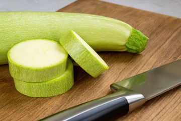 Close-up of fresh green zucchini, few slices and chef knife on a brown wooden cutting board. Cook at home. Fresh farm vegetables and healthy vegetarian food.