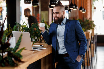 Young serious bearded businessman working on computer at table,drinking coffee.Man analyzes information, data, develops business plan. Freelancer, entrepreneur.Online marketing, education, e-learning