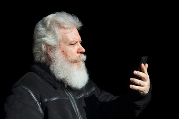Handsome senior man looking at smartphone against black background. Close up profile portrait of elderly stylish male with splendid mustache and beard wearing dark flight (bomber) jacket.