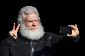 Handsome senior man looking at smartphone and showing a peace sign against nature background. Close up face portrait of elderly stylish male with splendid mustache and beard wearing dark flight jacket