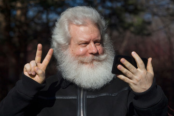 Cheerful senior man looking at smartphone and showing a peace sign against nature background. Close up face portrait of elderly stylish male with splendid mustache and beard wearing dark flight jacket