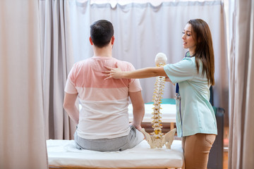 Beautiful female doctor with brown hair holding spine model and touching patient on his backs.