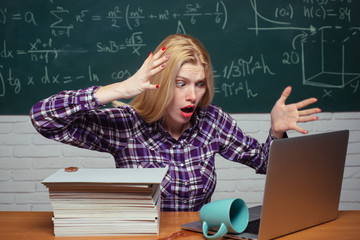 High school. Young teacher near chalkboard in school classroom. Tired student. Student sitting at table and writing on notebook. Bored student doing homework on line with a laptop