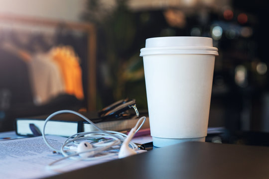 Closeup of a white paper cup of coffee on a table in an empty cafe without people. Nearby are the headphones and notebook. Mockup.