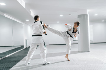 Fototapeta na wymiar Young Caucasian boy in dobok kicking barefoot while trainer holding kick target. Taekwondo training concept.