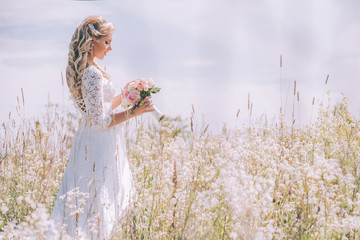 Bride in a white lace dress with an open back in a field of white flowers. The girl holds a delicate wedding bouquet of pink roses. Summer sun and blurred background.