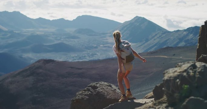Young adventurous woman leaps onto a rock in slow motion and enjoys the view from the top of a mountain with her arms raised in joy, amazing summer adventures