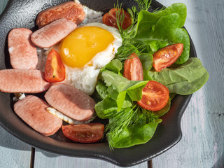 A rich breakfast of scrambled eggs with sausages, cherry tomatoes and leaf lettuce on a black round pan. Serve on a wooden plank tray with bread rolls as a reference fork.