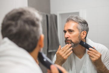 middle aged bearded gray haired man trimming his beard in bathroom