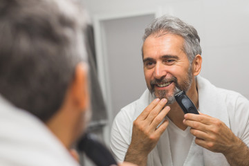 middle aged bearded gray haired man trimming his beard in bathroom