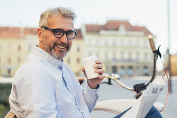 happy senior man reading newspaper and drinking coffee to go outdoor in the city