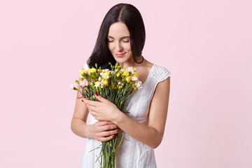 Indoor shot of brunette woman looks gently at flowers in her hands, dressed in white dress, has present from her boyfriend, posing isolated over rosy background. Copy space for promotion content.