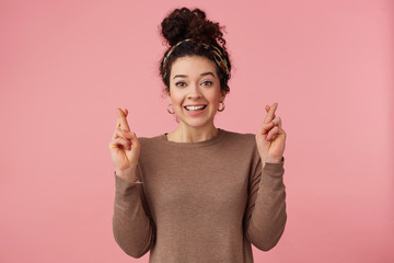 Portrait of happy young beautiful girl with curly dark hair, fingers crossed, hopes that she will be lucky, broadly smiling and looking at the camera isolated over pink background.