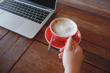 Selective focus of young female hand holding red cup of cappuccino coffee with blurred laptop computer on wooden tabletop in vintage tone style, close up with high angle view 
