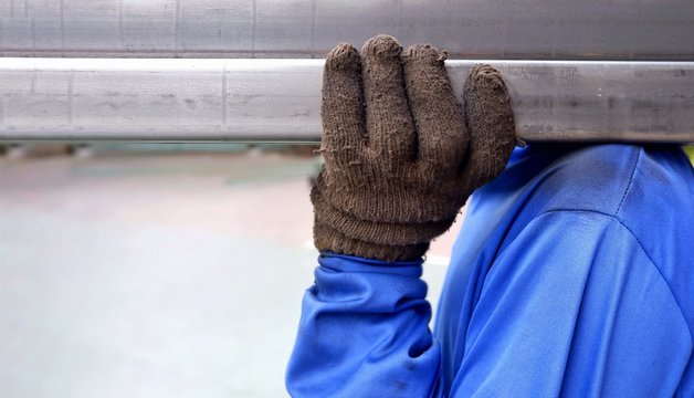 Close up worker man's hand in dirty woven glove carrying carbon steel rectangular tubes on his shoulder with blurred background, occupation concept
