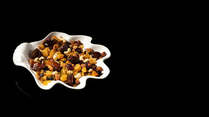 White plate of dried fruits on black background, with reflection. Mix of nuts and berries: raisins, hazelnut, cashews, almonds, cranberries, dried dates and apricots